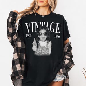 May include: Black t-shirt with the word "VINTAGE" above a black and white photo of a child wearing sunglasses and making a peace sign. The text "EST. 1996" is below the word "VINTAGE".