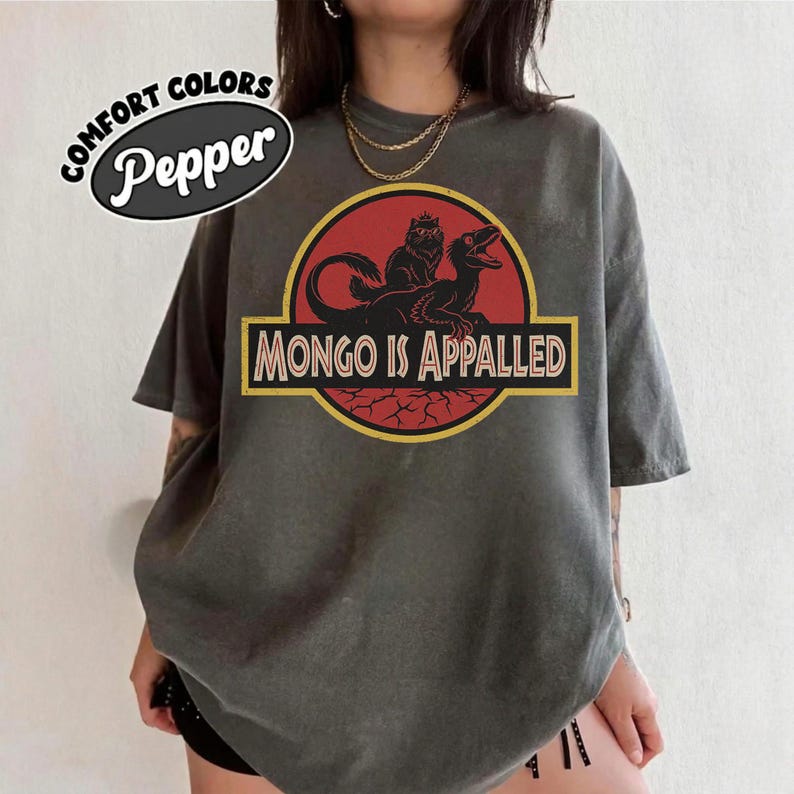 Peut inclure: T-shirt gris avec un motif de dinosaure et le texte "MONGO IS APPALLED" en jaune. Le motif est encadr&eacute; dans un cercle rouge et un rectangle jaune. Le t-shirt porte &eacute;galement le texte "COMFORT COLORS" et "Pepper" dans un ovale blanc.