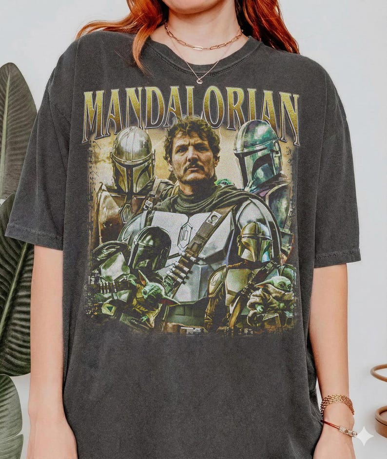K&ouml;nnte beinhalten: Dunkelgraues T-Shirt mit einer Grafik von Charakteren aus The Mandalorian. Das Design enth&auml;lt den Text "MANDALORIAN" in goldfarbenen Buchstaben &uuml;ber Bildern des Mandalorianers und anderer Charaktere in R&uuml;stung. Das T-Shirt hat einen Vintage-Look.
