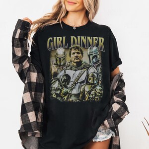 May include: Black t-shirt featuring a graphic of characters from a science fiction series. The text "GIRL DINNER" is printed above the graphic in a gold color. The graphic includes images of characters in armor.