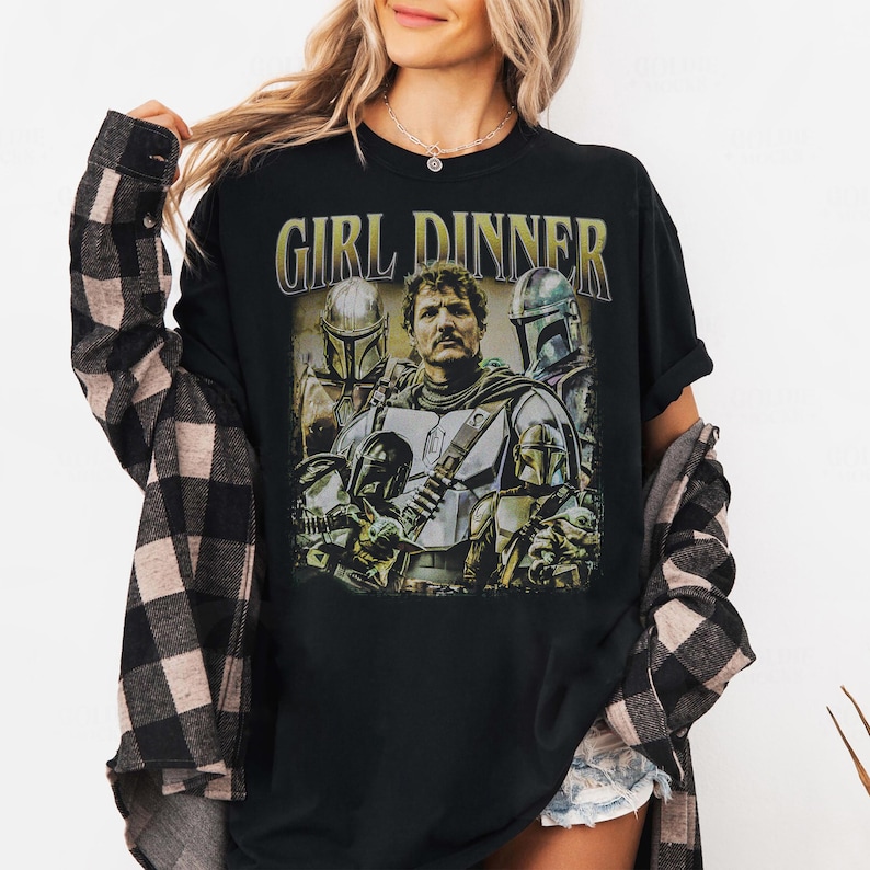 May include: Black t-shirt featuring a graphic of characters from a science fiction series. The text "GIRL DINNER" is printed above the graphic in a gold color. The graphic includes images of characters in armor.