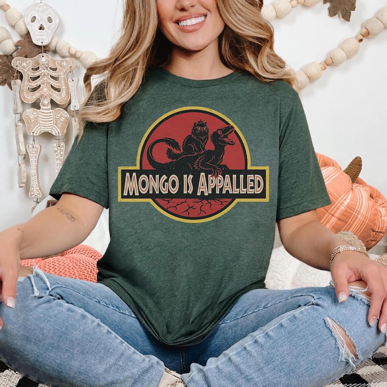 Peut inclure: T-shirt vert fonc&eacute; avec un motif repr&eacute;sentant un dinosaure et un chat, avec le texte "MONGO IS APPALLED". Le design est jaune et noir. Le t-shirt est &agrave; col rond et &agrave; manches courtes.