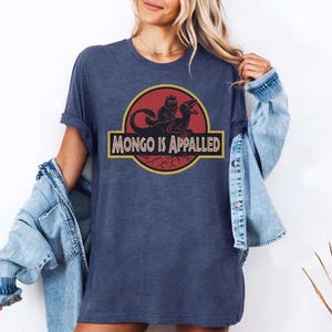 Peut inclure: T-shirt bleu marine avec un motif graphique repr&eacute;sentant un dinosaure et une cr&eacute;ature, avec le texte "MONGO IS APPALLED" dans un design jaune et noir. Le t-shirt est de style d&eacute;contract&eacute;, &agrave; manches courtes.