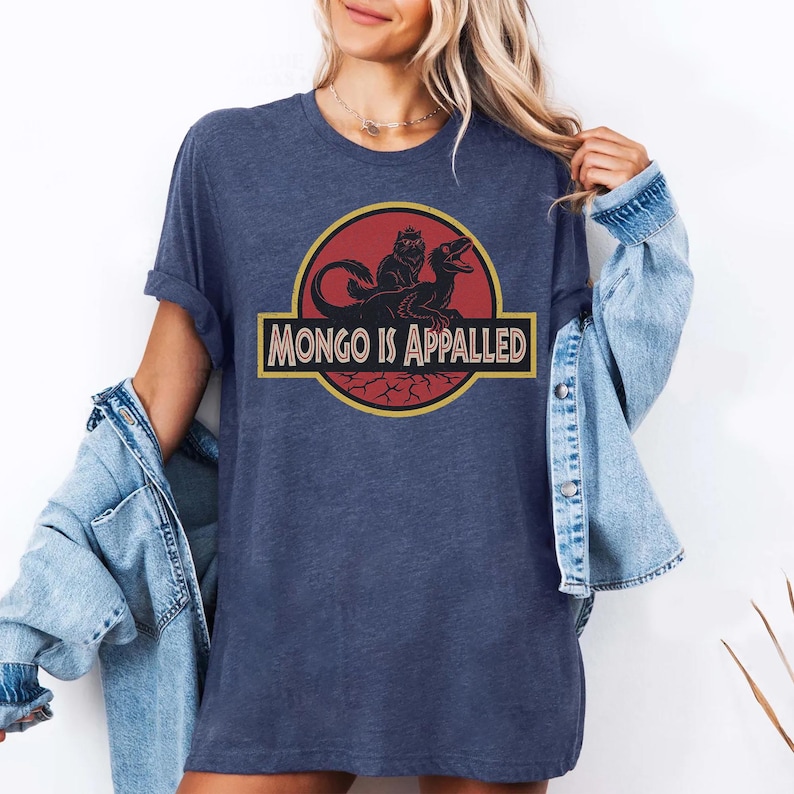 Peut inclure: T-shirt bleu marine avec un motif graphique repr&eacute;sentant un dinosaure et une cr&eacute;ature, avec le texte "MONGO IS APPALLED" dans un design jaune et noir. Le t-shirt est de style d&eacute;contract&eacute;, &agrave; manches courtes.