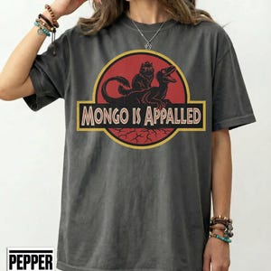 Peut inclure: T-shirt gris fonc&eacute; avec un motif repr&eacute;sentant un cercle rouge avec un dinosaure et un chat, et le texte "MONGO IS APPALLED" dans un rectangle jaune. Le t-shirt a des manches courtes et une coupe d&eacute;contract&eacute;e.