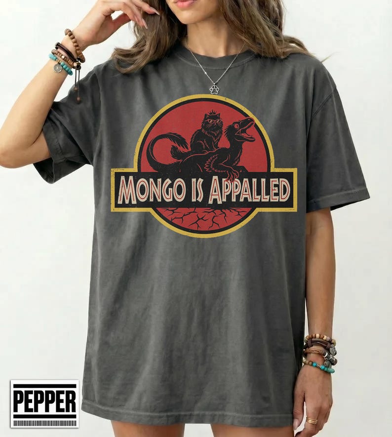 Peut inclure: T-shirt gris fonc&eacute; avec un motif repr&eacute;sentant un cercle rouge avec un dinosaure et un chat, et le texte "MONGO IS APPALLED" dans un rectangle jaune. Le t-shirt a des manches courtes et une coupe d&eacute;contract&eacute;e.