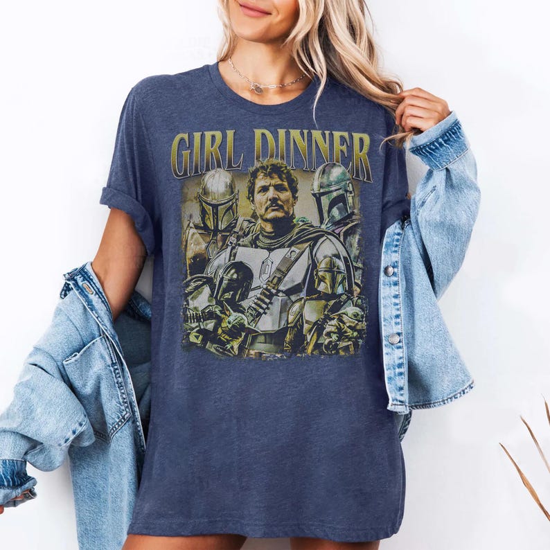 May include: A navy blue t-shirt featuring a graphic of characters from a science fiction series. The text "GIRL DINNER" is printed in gold above the image. The shirt is a relaxed fit.