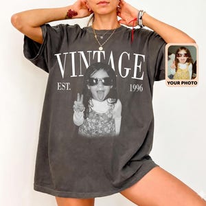 May include: Dark gray vintage t-shirt with the word "VINTAGE" in white block letters above a black and white photo of a child wearing sunglasses and making a peace sign. The text "EST. 1996" is below the word "VINTAGE". A small photo is on the right.