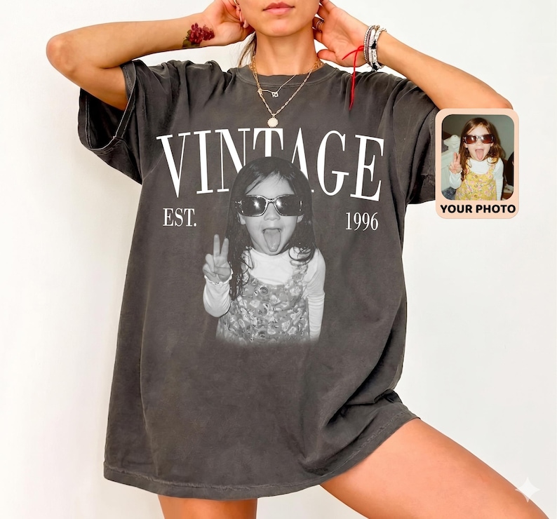 May include: Dark gray vintage t-shirt with the word "VINTAGE" in white block letters above a black and white photo of a child wearing sunglasses and making a peace sign. The text "EST. 1996" is below the word "VINTAGE". A small photo is on the right.
