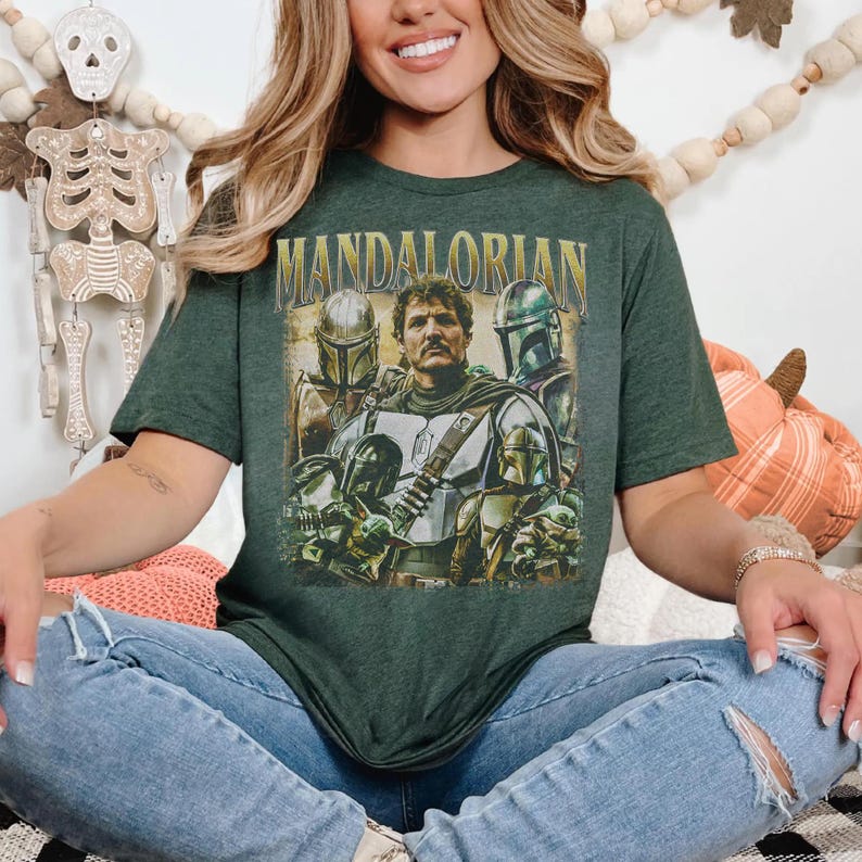 K&ouml;nnte beinhalten: Dunkelgr&uuml;nes T-Shirt mit dem Wort "MANDALORIAN" in goldfarbenen Buchstaben &uuml;ber einer Grafik von Charakteren aus der Star Wars-Serie. Die Grafik enth&auml;lt Bilder des Mandalorianers und anderer Charaktere in Helmen und R&uuml;stungen.