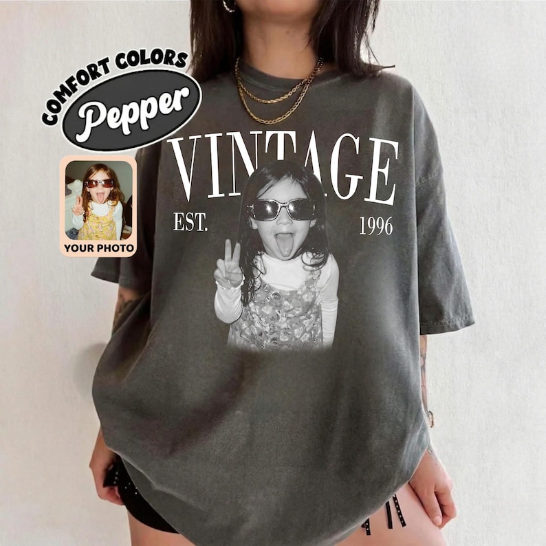 May include: Dark gray t-shirt with the word "VINTAGE" in white, along with "EST. 1996". A black and white photo of a child wearing sunglasses and making a peace sign is centered on the shirt. The shirt also has the words "COMFORT COLORS" and "Pepper" in a circle.