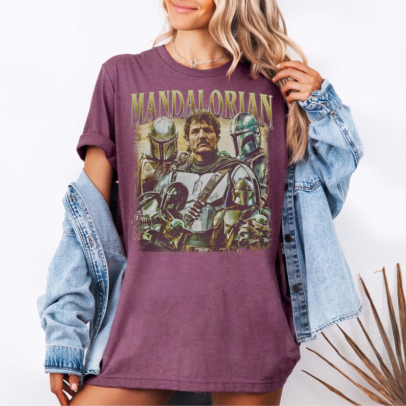 K&ouml;nnte beinhalten: Ein weinrotes T-Shirt mit einer Grafik von Charakteren aus der Mandalorian-Serie. Das Design enth&auml;lt den Text "MANDALORIAN" in goldfarbenen Buchstaben &uuml;ber einer Gruppe von Charakteren in R&uuml;stung. Das T-Shirt ist kurz&auml;rmlig und hat einen Rundhalsausschnitt.
