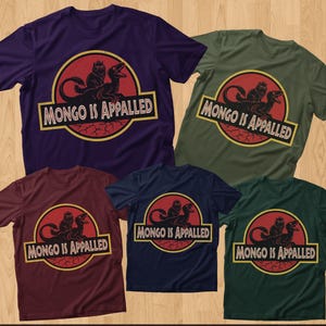 Peut inclure: Cinq t-shirts &agrave; manches courtes de diff&eacute;rentes couleurs : violet, vert olive, bordeaux, bleu marine et vert fonc&eacute;. Chaque chemise pr&eacute;sente un graphisme avec un dinosaure et le texte "MONGO IS APPALLED" dans un design jaune et rouge.