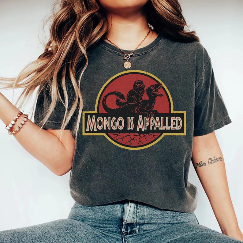 Peut inclure: T-shirt gris fonc&eacute; avec un graphisme humoristique. Le motif comprend un cercle rouge avec une silhouette noire d'une cr&eacute;ature et le texte "MONGO IS APPALLED" dans un rectangle bord&eacute; de jaune. V&ecirc;tement d&eacute;contract&eacute;.