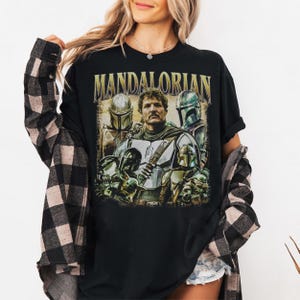 K&ouml;nnte beinhalten: Schwarzes T-Shirt mit einer Grafik des Mandalorianers und anderer Charaktere aus der Star Wars-Serie. Das Wort "MANDALORIAN" ist oben aufgedruckt. Das Design enth&auml;lt Bilder des Mandalorianers und anderer Charaktere mit Helmen.