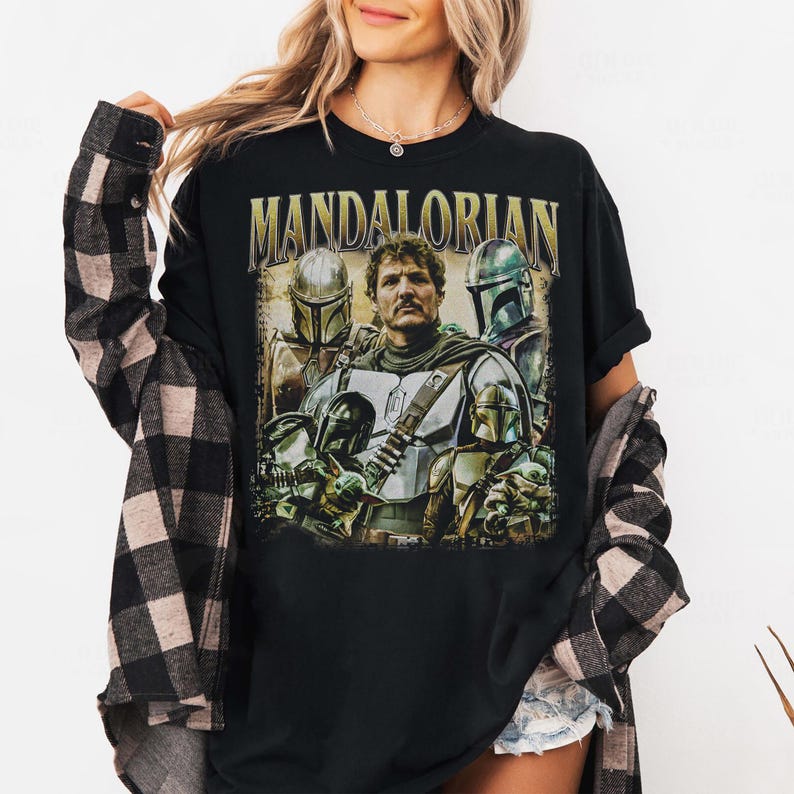 K&ouml;nnte beinhalten: Schwarzes T-Shirt mit einer Grafik des Mandalorianers und anderer Charaktere aus der Star Wars-Serie. Das Wort "MANDALORIAN" ist oben aufgedruckt. Das Design enth&auml;lt Bilder des Mandalorianers und anderer Charaktere mit Helmen.