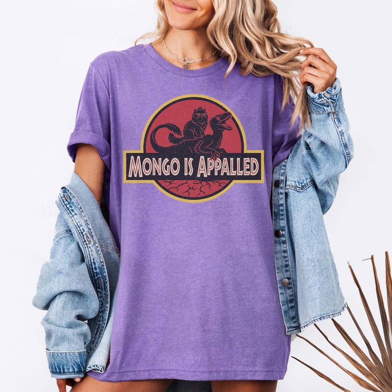 Peut inclure: Un t-shirt violet avec un motif de dinosaure et le texte "MONGO IS APPALLED" en jaune et noir. Le t-shirt est port&eacute; avec une veste en jean.
