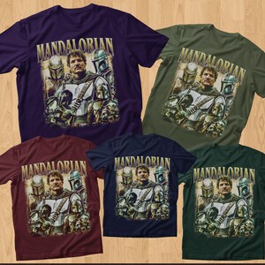 K&ouml;nnte beinhalten: F&uuml;nf kurz&auml;rmelige T-Shirts in verschiedenen Farben: Lila, Gr&uuml;n, Burgunderrot, Marineblau und Dunkelgr&uuml;n. Jedes Shirt zeigt eine Grafik eines Mannes mit Helmen und dem Wort "MANDALORIAN" in goldfarbenen Buchstaben. Die Shirts werden auf einer Holzoberfl&auml;che pr&auml;sentiert.