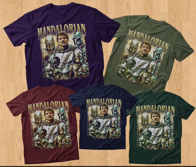 K&ouml;nnte beinhalten: F&uuml;nf kurz&auml;rmelige T-Shirts in verschiedenen Farben: Lila, Gr&uuml;n, Burgunderrot, Marineblau und Dunkelgr&uuml;n. Jedes Shirt zeigt eine Grafik eines Mannes mit Helmen und dem Wort "MANDALORIAN" in goldfarbenen Buchstaben. Die Shirts werden auf einer Holzoberfl&auml;che pr&auml;sentiert.