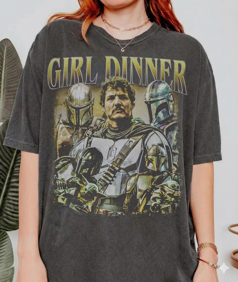 May include: Dark gray t-shirt featuring a graphic of characters from a science fiction series. The text "GIRL DINNER" is printed above the image. The image includes a central figure and several figures in helmets.