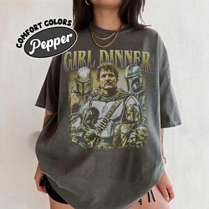 May include: Dark gray t-shirt with a graphic featuring characters from a science fiction series. The text "GIRL DINNER" is printed in yellow above the image. The shirt has a distressed look and the words "COMFORT COLORS" and "Pepper" are printed on the upper left.