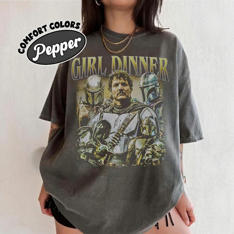 May include: Dark gray t-shirt with a graphic featuring characters from a science fiction series. The text "GIRL DINNER" is printed in yellow above the image. The shirt has a distressed look and the words "COMFORT COLORS" and "Pepper" are printed on the upper left.