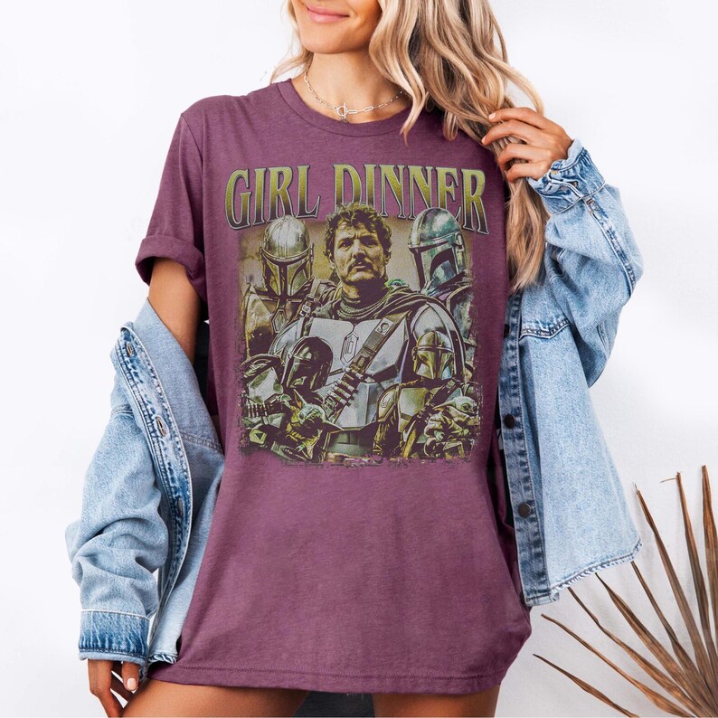 May include: A maroon t-shirt with the text "GIRL DINNER" above a graphic of a man and several figures in armor. The graphic has a vintage, distressed look. The shirt is a solid color and has short sleeves.
