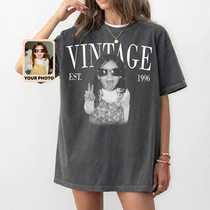 May include: Dark gray t-shirt with the word "VINTAGE" in white block letters above a black and white photo of a child wearing sunglasses and making a peace sign. The text "EST. 1996" is below the word "VINTAGE". A small photo is on the left side of the shirt.