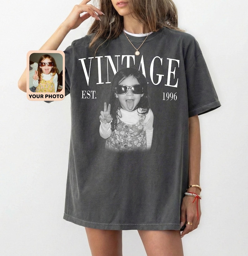 May include: Dark gray t-shirt with the word "VINTAGE" in white block letters above a black and white photo of a child wearing sunglasses and making a peace sign. The text "EST. 1996" is below the word "VINTAGE". A small photo is on the left side of the shirt.