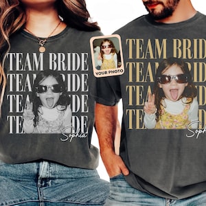 May include: Two charcoal gray t-shirts featuring a repeating white and gold text graphic that reads TEAM BRIDE. Each shirt displays a central photograph of a child wearing sunglasses and making a peace sign, with the name Sophia written below.