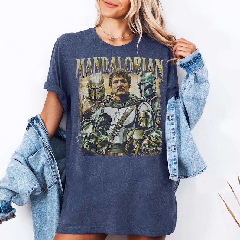 K&ouml;nnte beinhalten: Marineblaues T-Shirt mit einem Grafikdesign im Vintage-Stil des Mandalorianers. Das Design enth&auml;lt den Text "MANDALORIAN" in goldfarbenen Buchstaben sowie Bilder des Mandalorianers und anderer Charaktere aus der Serie.