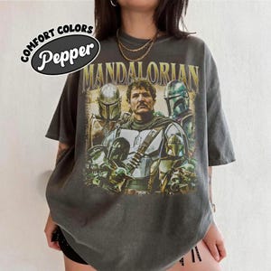 K&ouml;nnte beinhalten: Dunkelgraues T-Shirt mit einer Grafik von Charakteren aus The Mandalorian. Das Design enth&auml;lt den Text "MANDALORIAN" in goldfarbenen Buchstaben und das Logo der Marke Comfort Colors mit dem Wort "Pepper".