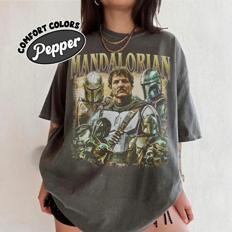 K&ouml;nnte beinhalten: Dunkelgraues T-Shirt mit einer Grafik von Charakteren aus The Mandalorian. Das Design enth&auml;lt den Text "MANDALORIAN" in goldfarbenen Buchstaben und das Logo der Marke Comfort Colors mit dem Wort "Pepper".