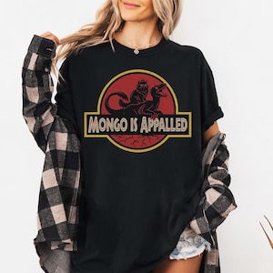 Peut inclure: T-shirt noir avec un motif rouge et jaune. Le motif repr&eacute;sente un dinosaure et une cr&eacute;ature avec le texte "MONGO IS APPALLED" en jaune. Le t-shirt est &agrave; col rond et &agrave; manches courtes.