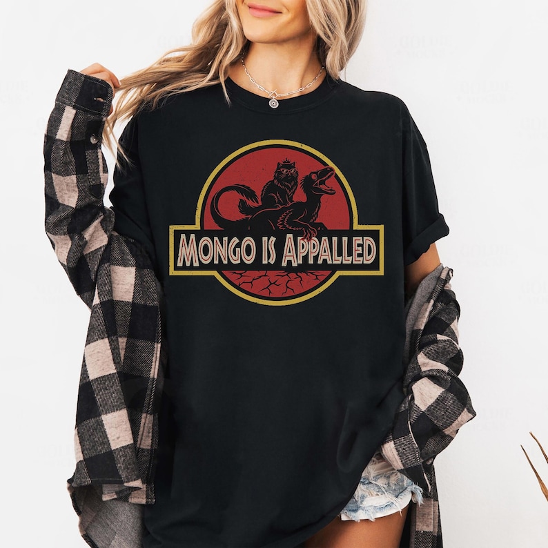 Peut inclure: T-shirt noir avec un motif rouge et jaune. Le motif repr&eacute;sente un dinosaure et une cr&eacute;ature avec le texte "MONGO IS APPALLED" en jaune. Le t-shirt est &agrave; col rond et &agrave; manches courtes.