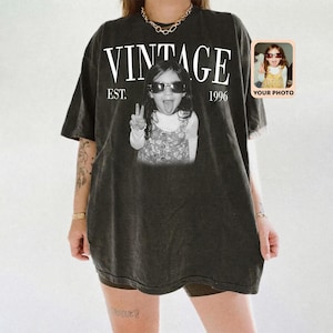 May include: Dark gray vintage-style t-shirt with the word "VINTAGE" in white block letters above a black and white photo of a child wearing sunglasses and making a peace sign. The text "EST. 1996" is below the word "VINTAGE". A small photo is in the upper right corner.