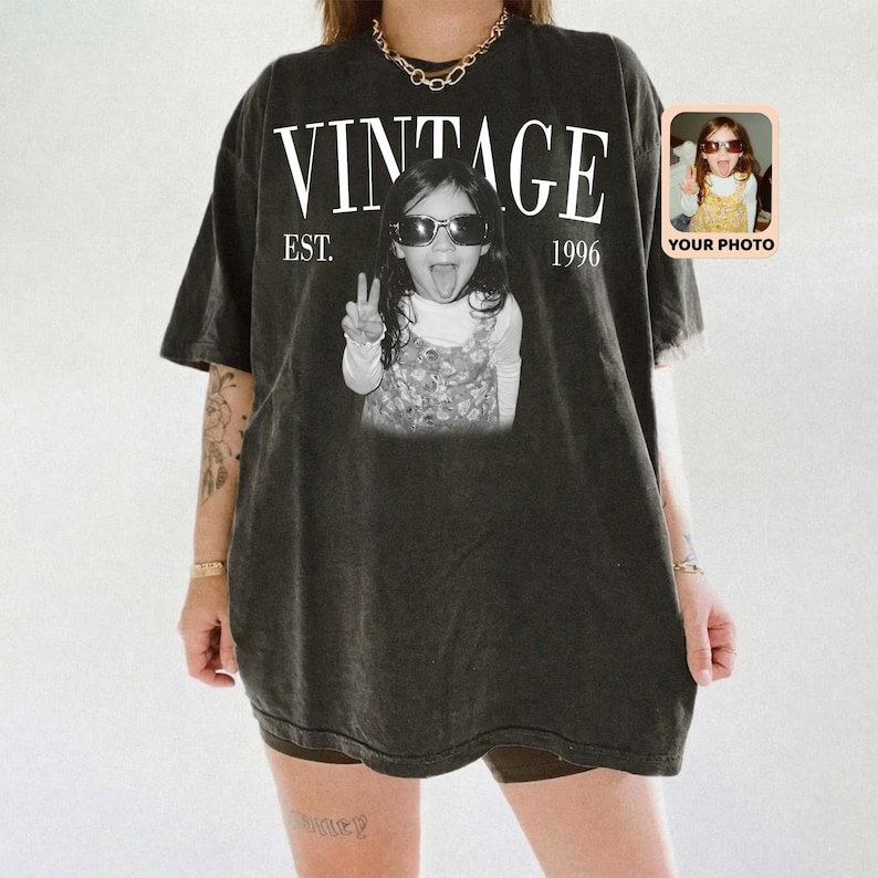May include: Dark gray vintage-style t-shirt with the word "VINTAGE" in white block letters above a black and white photo of a child wearing sunglasses and making a peace sign. The text "EST. 1996" is below the word "VINTAGE". A small photo is in the upper right corner.