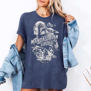 May include: Navy blue t-shirt featuring a graphic from The Mandalorian series. The design includes the Mandalorian helmet, Grogu, and the text "THE MANDALORIAN AND GROGU" and "THIS IS THE WAY" in white. The shirt has short sleeves.