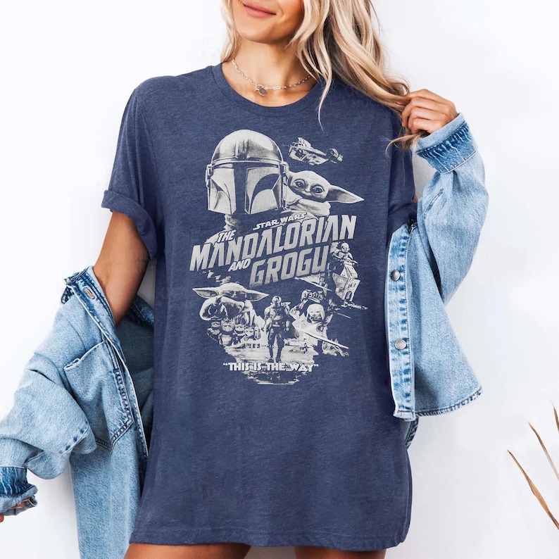May include: Navy blue t-shirt featuring a graphic from The Mandalorian series. The design includes the Mandalorian helmet, Grogu, and the text "THE MANDALORIAN AND GROGU" and "THIS IS THE WAY" in white. The shirt has short sleeves.