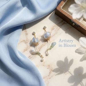 May include: A pair of floral earrings with a light blue flower design. The earrings have gold-coloured wire detailing, a clear gem, and a pearl drop. The image includes the text "Artistry in Bloom".