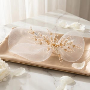 May include: A delicate ivory headpiece with a gold floral design and pearl accents. The headpiece is made of sheer fabric and is set on a gold-toned base. The design includes small, round pearls and gold-colored leaves.