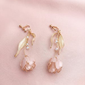 May include: A pair of clip-on earrings featuring a floral design. The earrings have a gold-colored metal frame with a cream-colored leaf and a light pink flower. Clear beads dangle from the leaf and flower.