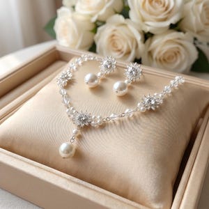 May include: A jewellery set comprising a necklace and earrings. The necklace and earrings are decorated with clear crystals, small pearls, and larger pearl drops. The set is presented on a beige satin cushion, with white roses in the background.