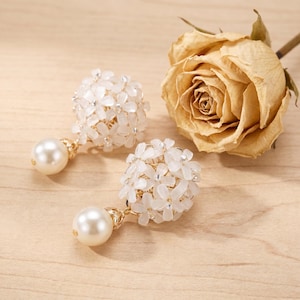 Peut inclure: Une paire de boucles d'oreilles dorées composées de grappes sphériques de perles blanches translucides en forme de fleurs, chacune agrémentée d'une perle nacrée crème suspendue. Les perles florales sont disposées en une forme arrondie dense.