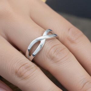 Peut inclure: Une bague argentée avec un motif de symbole infini. La bague présente deux bandes incurvées qui s'entrecroisent pour créer une forme ouverte et fluide. La bague est portée sur un doigt, mettant en valeur sa surface polie et réfléchissante.