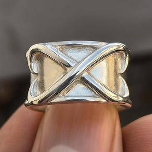 Könnte beinhalten: Ein silberner Ring mit einem X-förmigen Design. Der Ring hat ein breites Band, wobei sich das X-Muster um den Umfang legt. Der Ring wird von den Fingern einer Person gehalten, was seine polierte Oberfläche und die komplizierten Details hervorhebt.