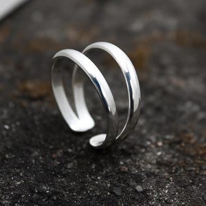 May include: A shiny silver double-band ring. The ring has a smooth, polished surface and a minimalist design. The bands are parallel and slightly curved, creating an open, modern look. The ring is set against a dark, textured background.