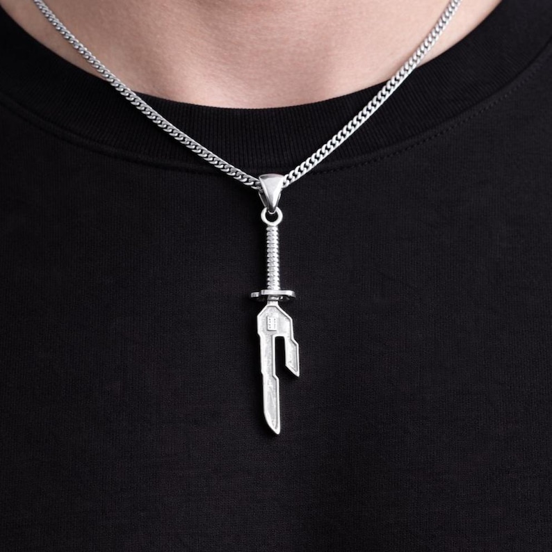May include: Silver-colored pendant necklace featuring a sword-shaped charm. The charm has a detailed design with a handle and blade. The necklace chain is a curb style. The pendant hangs against a black background.