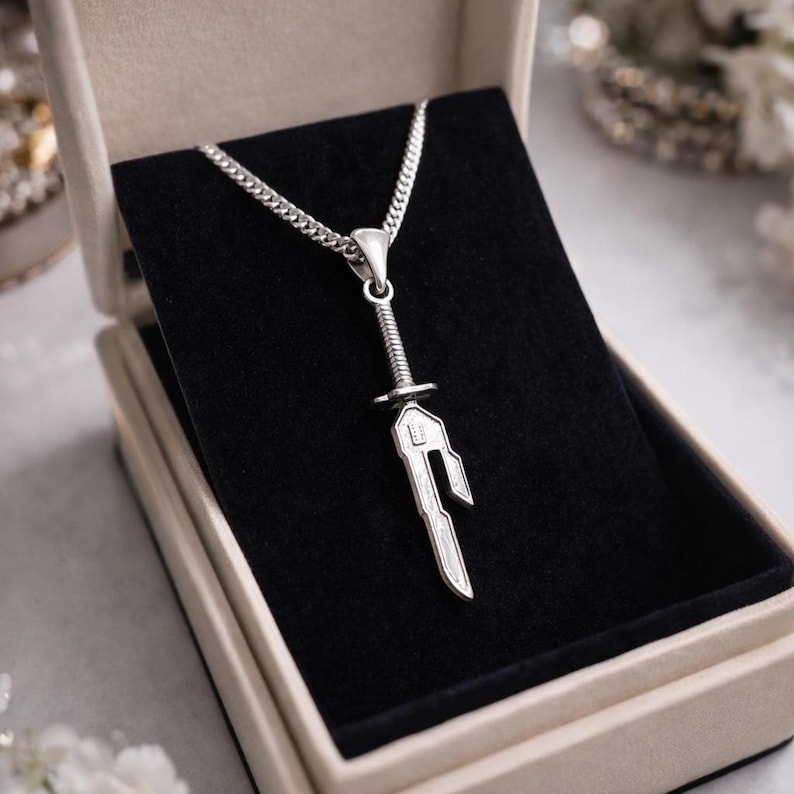 May include: A silver-colored pendant necklace featuring a sword design. The sword pendant is detailed with a hilt, blade, and decorative elements. The necklace is presented in a beige jewelry box with a black velvet interior.