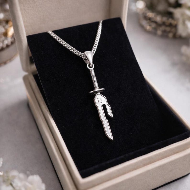 May include: A silver-colored pendant necklace featuring a sword design. The sword pendant is detailed with a hilt, blade, and decorative elements. The necklace is presented in a beige jewelry box with a black velvet interior, creating a striking contrast.